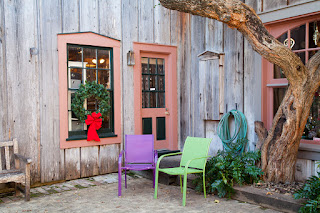 A corner shop with pastel colored trim has purple and green chairs out front, including a holiday wreath, in Salado, Texas