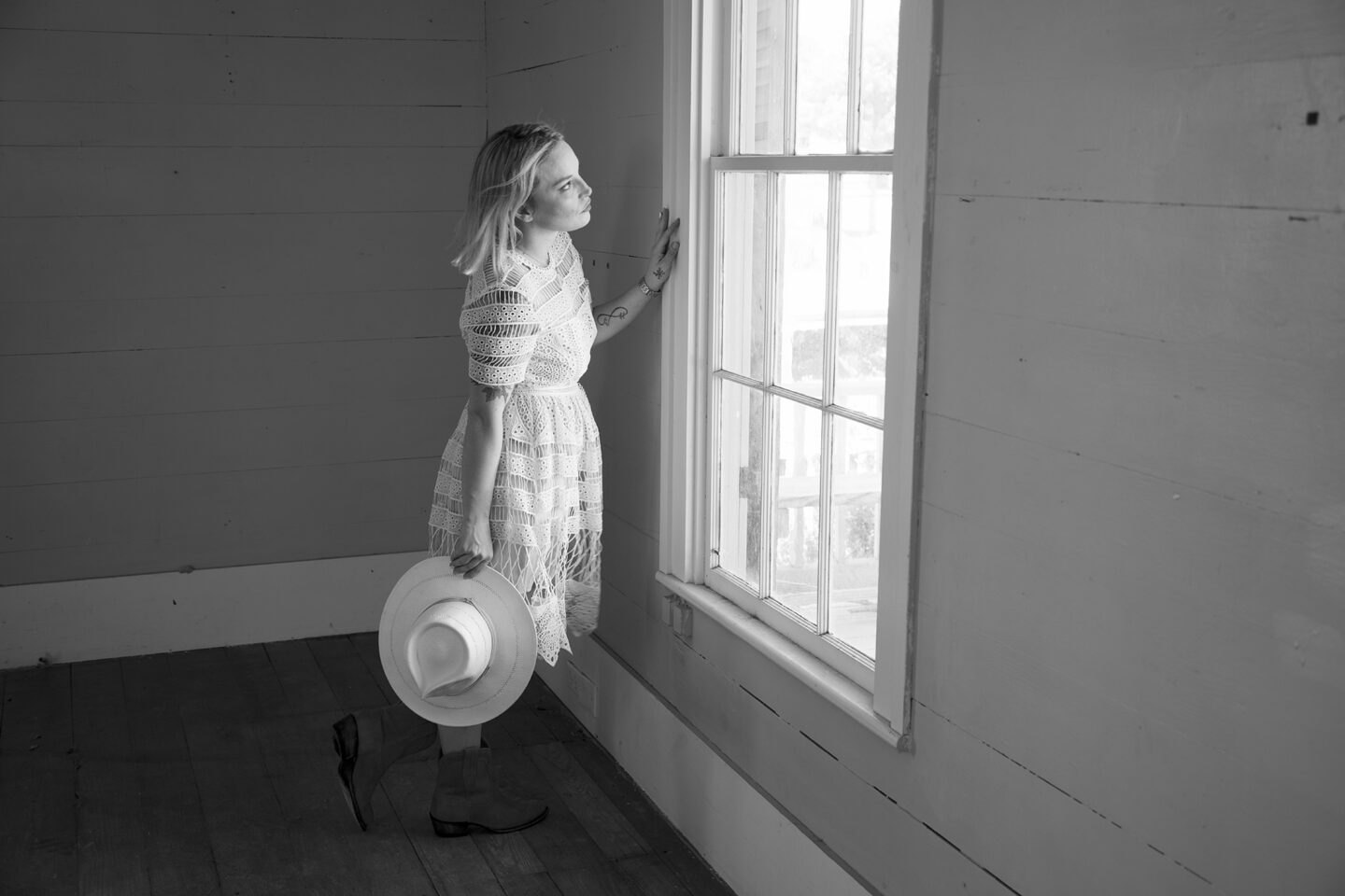Black and white image, portrait of a woman in a dress holding a cowboy hat gazing out a window 
