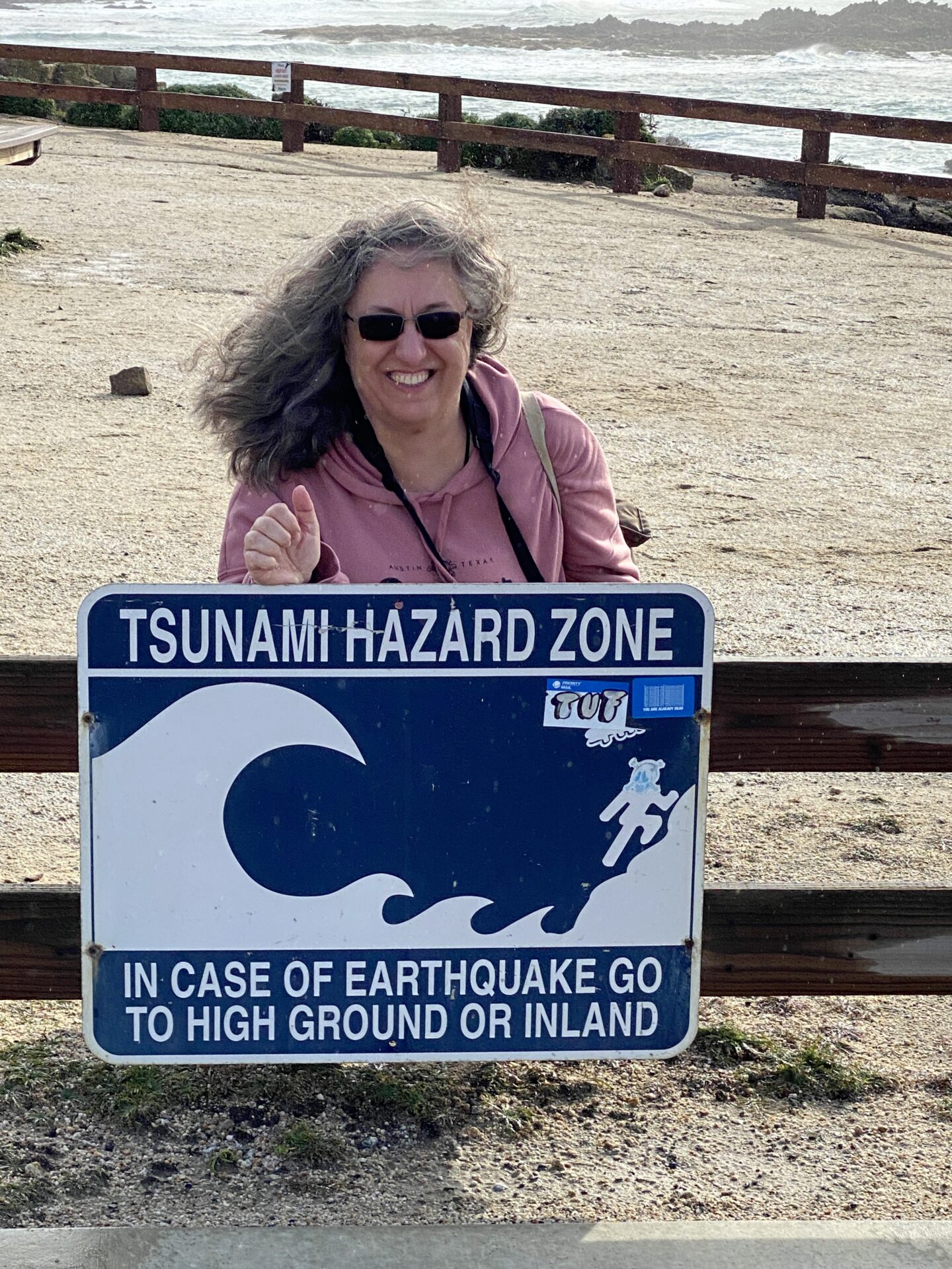 Portrait of Carol Schiraldi at the beach standing behind a sign that reads Tsunami Hazard Zone in the wind. 