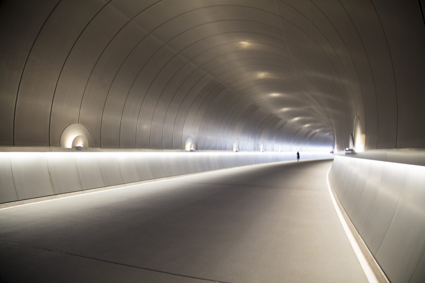 A shadow figure walks through the end of a long curved tunnel that is a liminal space included in the Out of Time/Out of Place exhibition by Carol Schiraldi.